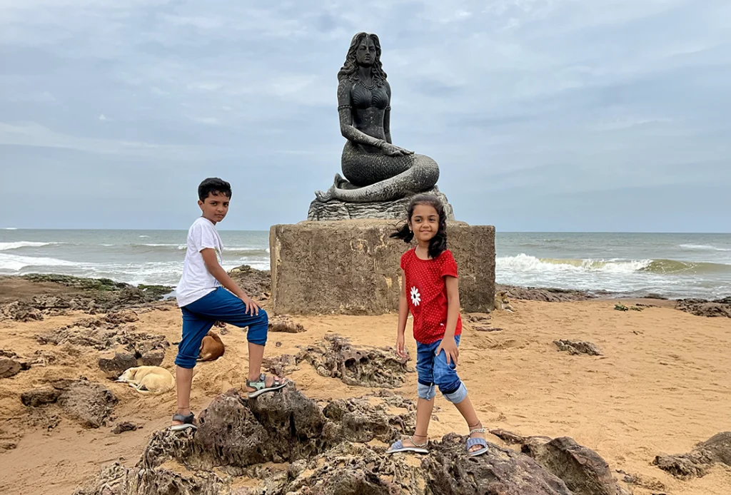 A white t shirt blue jeans boy and a girl with red shirt and blue jeans  standing in front of mermaid statue at bheemili beach